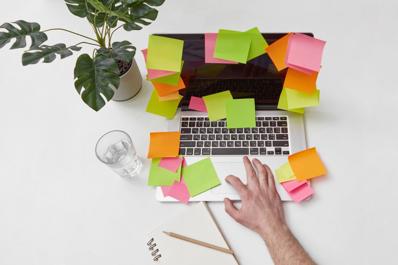 Laptop billentyűzeten kéz, körülötte színes post-it cetlik, mellette növény és jegyzetfüzet.